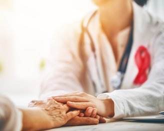 Eine Ärztin hält die Hand eines Patienten, während sie ihm aufmerksam zuhört. Im Hintergrund ist ein Stethoskop sichtbar.