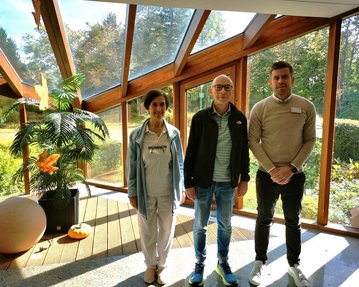 Drei Personen stehen in einem lichtdurchfluteten Raum mit großen Fenstern. Im Hintergrund sind Pflanzen und eine grüne Landschaft sichtbar.