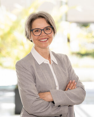 Eine lächelnde Frau mit grauen Haaren und Brille steht mit verschränkten Armen in einem modernen, hellen Raum.
