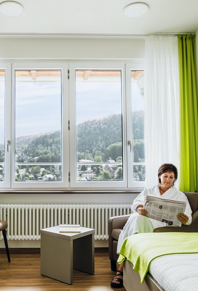 Eine Person in einem Bademantel sitzt auf einem Sofa in einem hellen Zimmer, liest eine Zeitung und blickt aus dem Fenster auf die Landschaft.