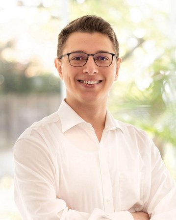 Ein junger Mann mit Brille trägt ein weißes Hemd und lächelt. Er steht mit verschränkten Armen vor einem hellen Hintergrund mit Pflanzen. Natürliches Licht strömt durch die Fenster.