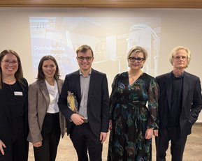 Fünf Personen stehen in einem modernen Raum, lächelnd und in formeller Kleidung. Im Hintergrund ist ein Präsentationsbild mit dem Text "Durchdachte Ganztagesangebote: Inklusion".