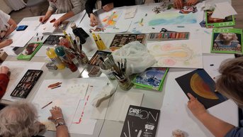Mehrere Personen arbeiten an verschiedenen Kunstprojekten an einem Tisch, umgeben von Farben, Stiften und Papier.