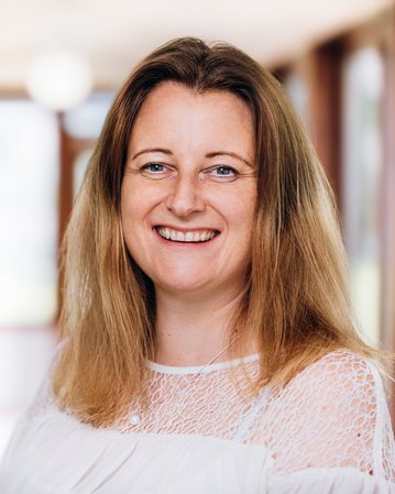 Eine Frau mit langen, blonden Haaren lächelt in einem hellen Flur. Sie trägt ein weißes Oberteil und steht leicht seitlich zur Kamera. Der Hintergrund zeigt unscharfe Fenster und eine helle, einladende Atmosphäre.