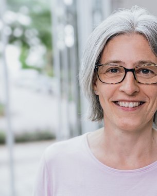 Eine lächelnde Frau mit grauen Haaren und Brille trägt ein helles Oberteil. Sie steht in einem modernen, hellen Raum mit unscharfem Hintergrund.