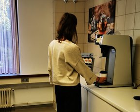 Eine Person steht an einer Kaffeemaschine in einer hellen Küche und füllt einen Becher mit Kaffee. Fenster mit Vorhängen im Hintergrund.