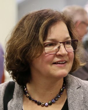 Eine Frau mit lockigem, braunem Haar und Brille trägt eine bunte Halskette und lächelt leicht. Sie ist in einem Innenraum mit unscharfen Personen im Hintergrund.