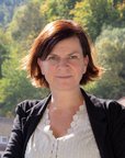 Eine Frau mit schulterlangen, braunen Haaren steht im Freien, vor einer bewaldeten Landschaft mit bunten Herbstblättern. Sie trägt eine schwarze Jacke und ein weißes Oberteil.