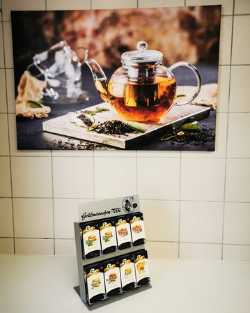 Teekanne mit schwarzem Tee auf einem Holzbrett, im Hintergrund ein dekoratives Bild. Vorne ein Ständer mit verschiedenen Teesorten.
