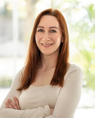 Eine lächelnde Frau mit langen, braunen Haaren steht mit verschränkten Armen vor einem hellen Hintergrund. Sie trägt ein helles Oberteil und hat eine freundliche Ausstrahlung. Im Hintergrund sind Pflanzen sichtbar.