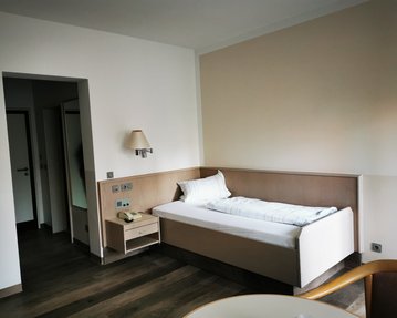 Ein einfaches Zimmer mit einem Einzelbett, Nachttisch, Lampe und einem Tisch. Im Hintergrund sind Türen zu weiteren Räumen sichtbar.