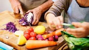 Zwei Personen bereiten frisches Gemüse vor, darunter Rotkohl, Karotten, Tomaten und Zitrone, während sie in einer Küche arbeiten.
