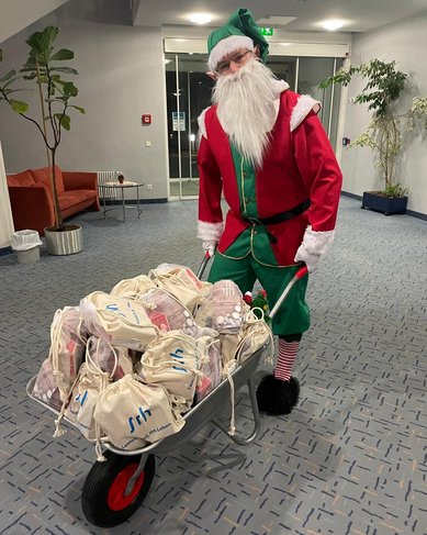Ein Weihnachtsmann in einem roten Kostüm schiebt eine Schubkarre voller Geschenktüten durch einen Innenraum.