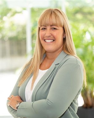 Frau mit langen blonden Haaren, die in einem hellen Blazer lächelt und mit verschränkten Armen vor einem hellen Hintergrund steht.