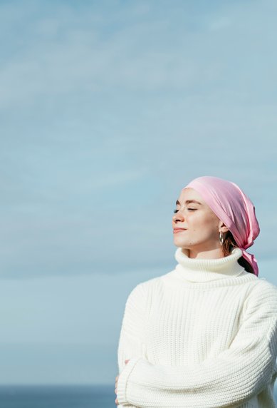 Eine Frau mit einem rosa Kopftuch und einem weißen Pullover steht am Meer und blickt entspannt in die Ferne.