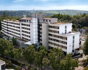 Ein mehrstöckiges Wohngebäude mit einer modernen Fassade, umgeben von Bäumen und Grünflächen. Im Hintergrund sind sanfte Hügel sichtbar.