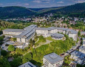 Ein modernes Gebäudeensemble in einer bewaldeten Umgebung, umgeben von Hügeln und einer kleinen Stadt im Hintergrund. Die Architektur ist vielfältig, mit mehreren Gebäuden und Grünflächen.