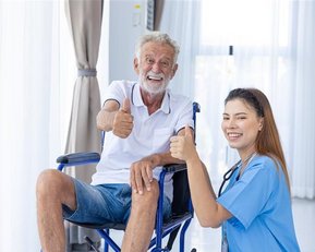 Ein älterer Mann im Rollstuhl und eine junge Frau geben beide Daumen hoch, lächelnd in einem hellen Raum.