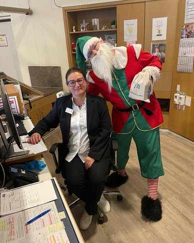 Eine Frau in einem schwarzen Blazer sitzt an einem Schreibtisch, während ein Weihnachtsmann hinter ihr steht und lächelt.