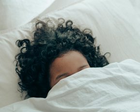 Ein Kind mit lockigem Haar liegt im Bett und ist teilweise mit einer Decke zugedeckt.