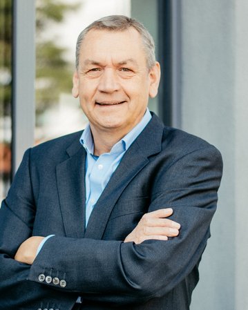 Ein Mann mittleren Alters steht mit verschränkten Armen vor einem modernen Gebäude. Er trägt einen dunkelblauen Anzug und ein hellblaues Hemd, mit einem neutralen Ausdruck im Gesicht.
