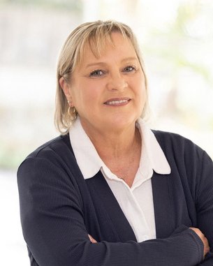 Eine Frau mit blonden Haaren steht mit verschränkten Armen in einem hellen Raum. Sie trägt ein dunkles Oberteil und ein weißes Hemd. Im Hintergrund sind unscharfe Pflanzen sichtbar.