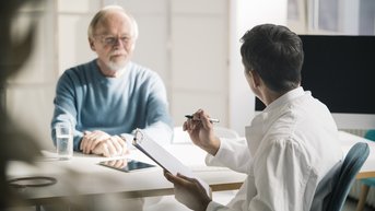 Ein älterer Mann sitzt an einem Tisch und schaut einem jüngeren Mann gegenüber, der in einem weißen Hemd sitzt und Notizen macht. Auf dem Tisch stehen ein Glas Wasser und ein Smartphone.