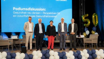 Podiumsdiskussion mit fünf Personen, die über Gesundheit und Zukunftsperspektiven sprechen. Im Hintergrund ein Banner mit dem Thema der Diskussion.