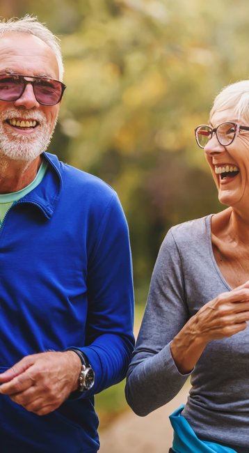 Ein älteres Paar joggt lächelnd auf einem Waldweg, beide tragen sportliche Kleidung und Sonnenbrillen.