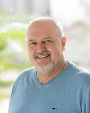 Ein lächelnder Mann mit einem grauen Bart trägt einen blauen Pullover. Er steht vor einer hellen, unscharfen Hintergrundlandschaft mit Pflanzen.