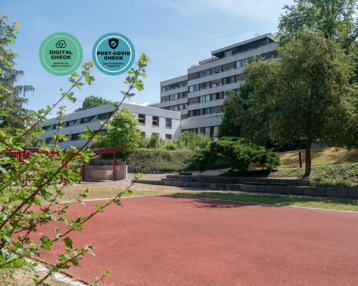 Ein roter Sportplatz mit einem modernen Gebäude im Hintergrund, umgeben von Bäumen. Zwei Symbole für digitale und Post-COVID-Checks sind sichtbar.