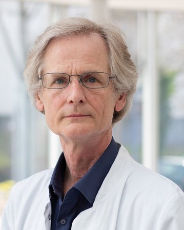 Ein älterer Mann mit grauen Haaren und Brille trägt einen weißen Kittel. Er schaut direkt in die Kamera, im Hintergrund sind Fenster sichtbar.