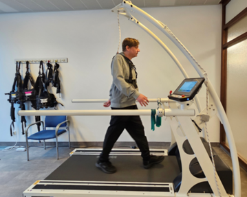 Ein Mann läuft auf einem speziellen Laufband mit Haltegestell in einem Trainingsraum. Ein Bildschirm zeigt Trainingsdaten an.