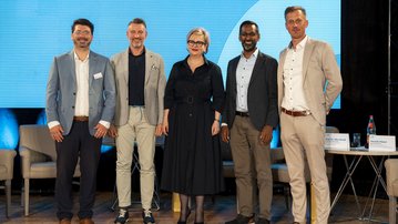Fünf Personen stehen nebeneinander auf einer Bühne, lächelnd, in formeller Kleidung, vor einem blauen Hintergrund mit Scheinwerferlicht.