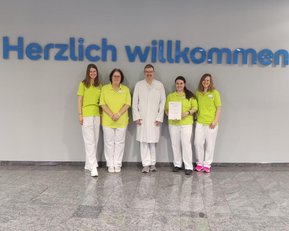 Fünf Personen in Arbeitskleidung stehen vor einer Wand mit der Aufschrift "Herzlich willkommen" und halten ein Zertifikat.