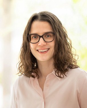 Eine Frau mit lockigem, braunem Haar und Brille lächelt in die Kamera. Sie trägt ein helles Hemd und steht vor einer hellen, unscharfen Hintergrundszene mit Pflanzen.