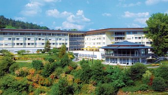 Ein modernes Hotelgebäude mit mehreren Etagen, umgeben von grünen Gärten und Bäumen. Im Vordergrund befindet sich ein Pavillon mit einer überdachten Terrasse. Der Himmel ist klar mit einigen Wolken.