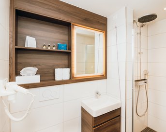 Ein modernes Badezimmer mit einer Dusche, einem Waschbecken und einem Holzregal. Auf dem Regal befinden sich Handtücher, Pflegeprodukte und ein Spiegel mit Beleuchtung.