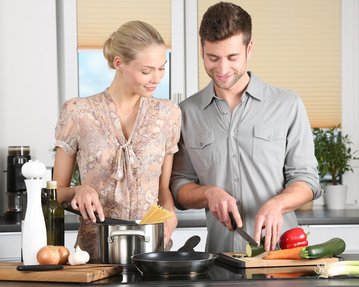 Ein Mann und eine Frau bereiten gemeinsam eine Mahlzeit in einer modernen Küche vor. Der Mann schneidet Gemüse, während die Frau Spaghetti in einem Topf hält. Auf der Arbeitsfläche sind verschiedene Zutaten und Kochutensilien sichtbar.