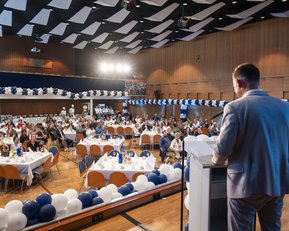 Ein Redner steht an einem Podium und spricht vor einem Publikum in einem festlich dekorierten Saal mit Tischen und Ballons.