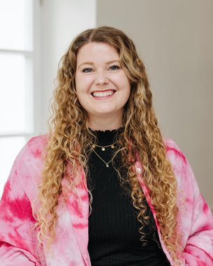 Eine lächelnde Frau mit langen, lockigen Haaren trägt einen pinken, gefärbten Mantel und steht in einem hellen Raum mit Fenstern.