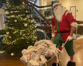 Ein Weihnachtsmann steht neben einem geschmückten Baum und schiebt eine Schubkarre voller Geschenke in einem festlichen Innenraum.
