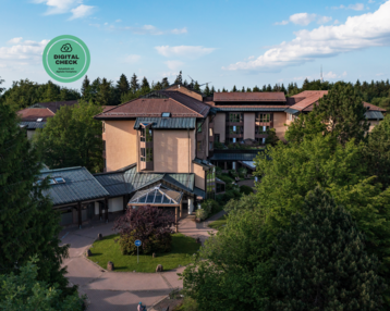 Ein mehrstöckiges Gebäude umgeben von Bäumen und Grünflächen, mit einem modernen Glasdach und einem digitalen Prüfsymbol in der oberen linken Ecke.