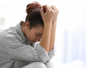 Eine Frau sitzt mit gesenktem Kopf und hält sich die Hände am Kopf, in einem Raum mit sanftem Licht.