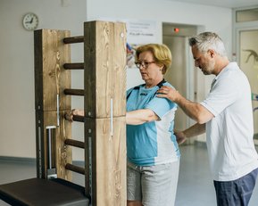 Eine Frau steht an einem Trainingsgerät, während ein Mann sie unterstützt. Beide sind in einem modernen Raum für Physiotherapie.