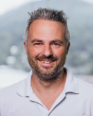 Ein Mann mit grauen Haaren und Bart lächelt in die Kamera. Er trägt ein weißes Polohemd und steht vor einer unscharfen, grünen Landschaft.