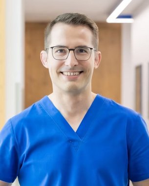 Ein Mann in einem blauen Arztkittel lächelt freundlich und steht in einem modernen, hellen Flur.