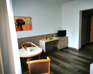 Ein heller Raum mit einem Tisch, Stühlen, einem Fernseher und einem Schreibtisch. An der Wand hängt ein Gemälde eines Hundes.