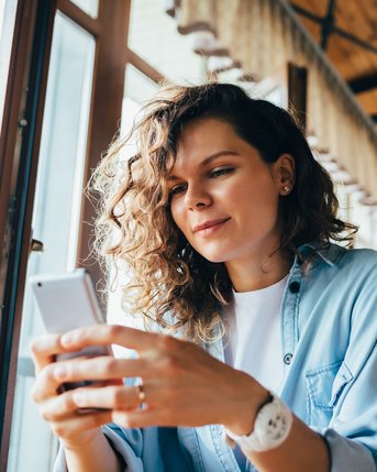 Eine Frau mit lockigem Haar sitzt in einem Café und schaut auf ihr Smartphone. Sie trägt ein lässiges Hemd und wirkt konzentriert.