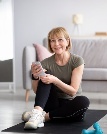 Eine lächelnde Frau sitzt auf einer Trainingsmatte in einem modernen Raum. Sie trägt sportliche Kleidung und hält ein Smartphone in der Hand. Neben ihr befinden sich eine Wasserflasche und ein Paar Hanteln.